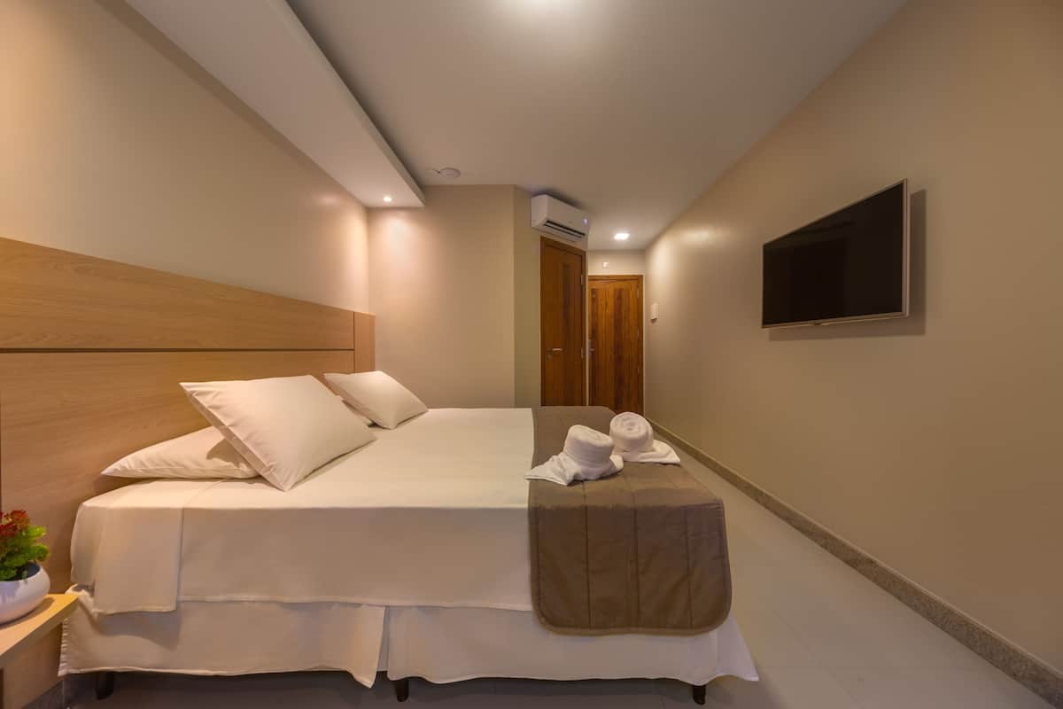 A hotel room with a bed, a TV mounted on the wall, a wooden headboard, and a small bedside table with a plant.