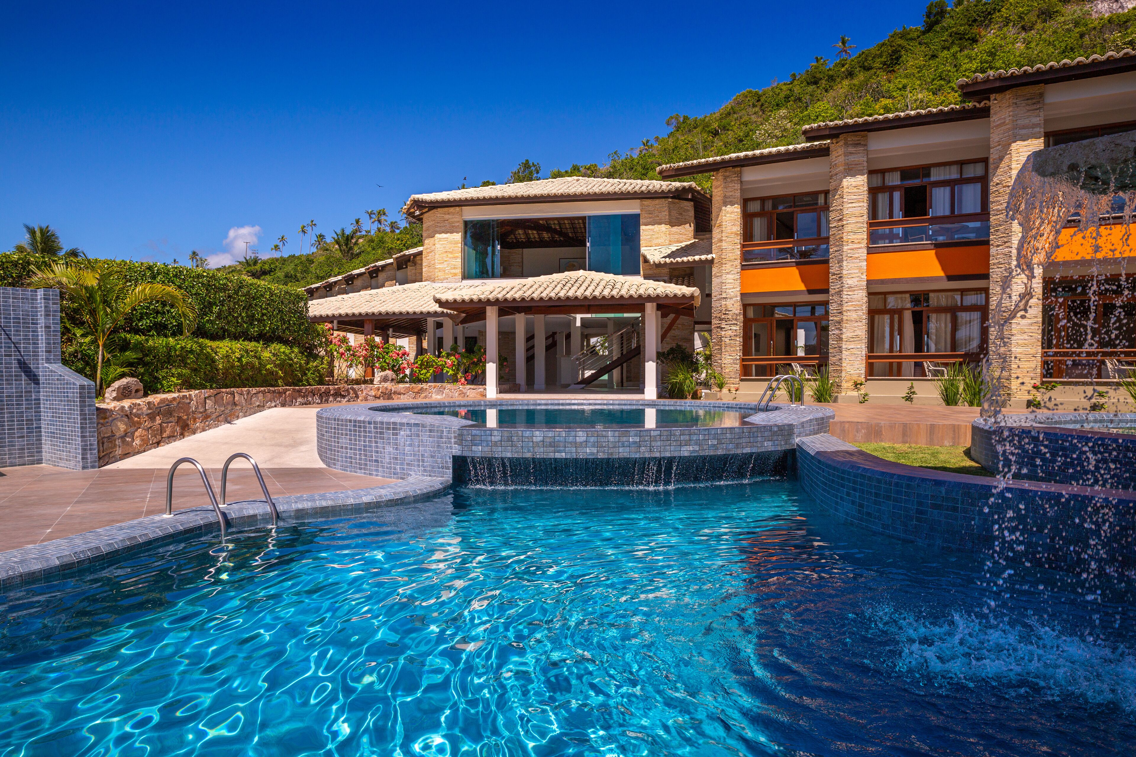 A swimming pool with a clear blue water feature, surrounded by a stone and tiled deck, and a multi-story building with balconies and a covered patio area.