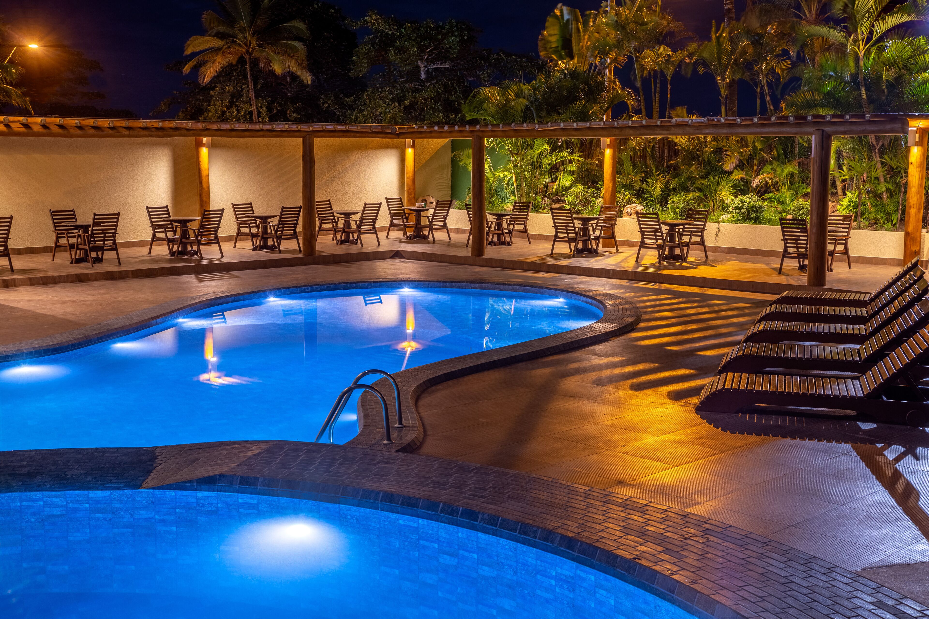 A pool area with lounge chairs and tables, surrounded by palm trees and a covered seating area.