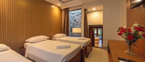 A hotel room with three single beds, a wooden desk with a vase of flowers, and a TV mounted on the wall.