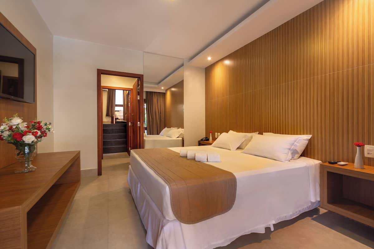 A modern hotel room with a large bed, a wooden headboard, a bedside table with a vase of flowers, and a mirror on the wall.