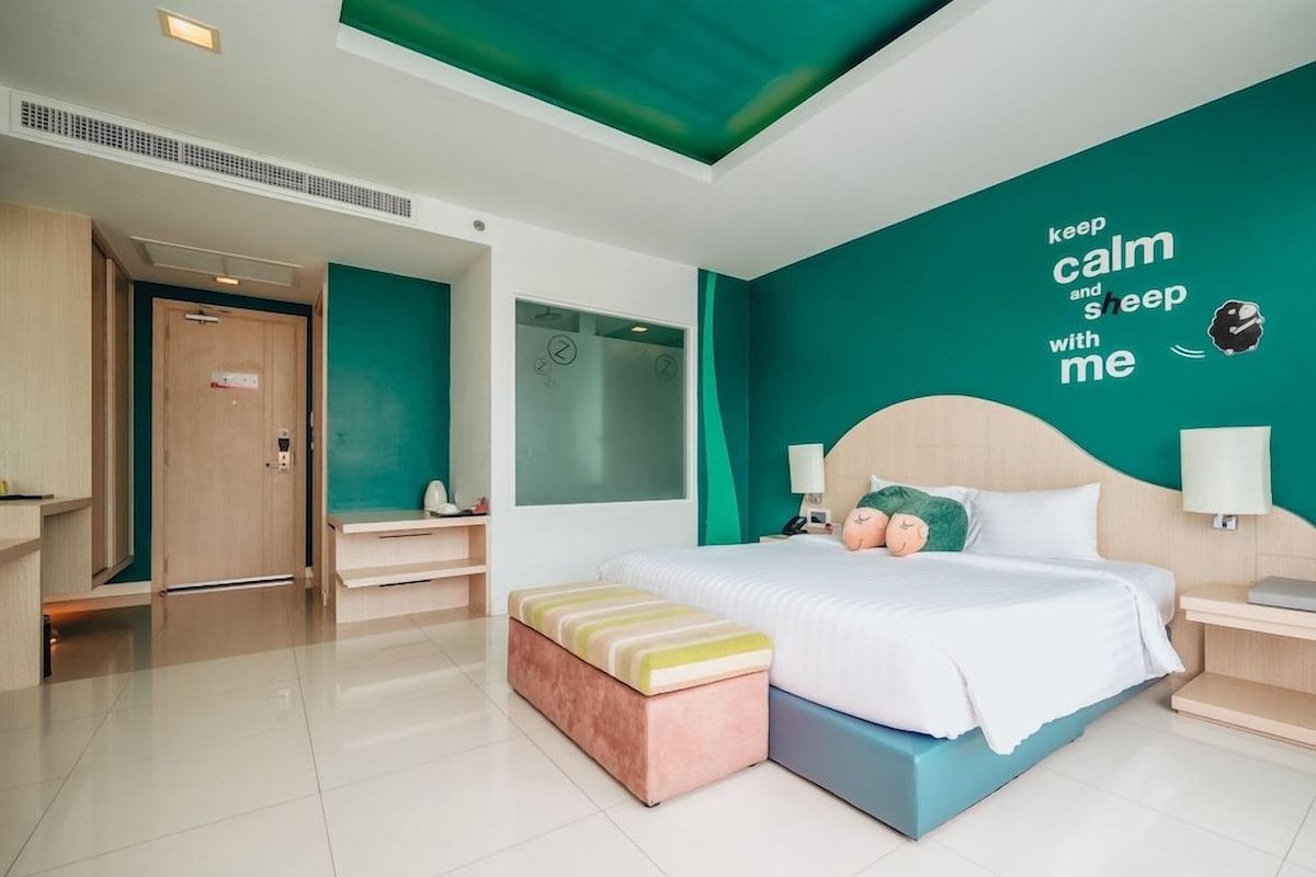 A modern hotel room with a large bed, a bedside table, a nightstand, a wall clock, and a motivational quote on the wall.