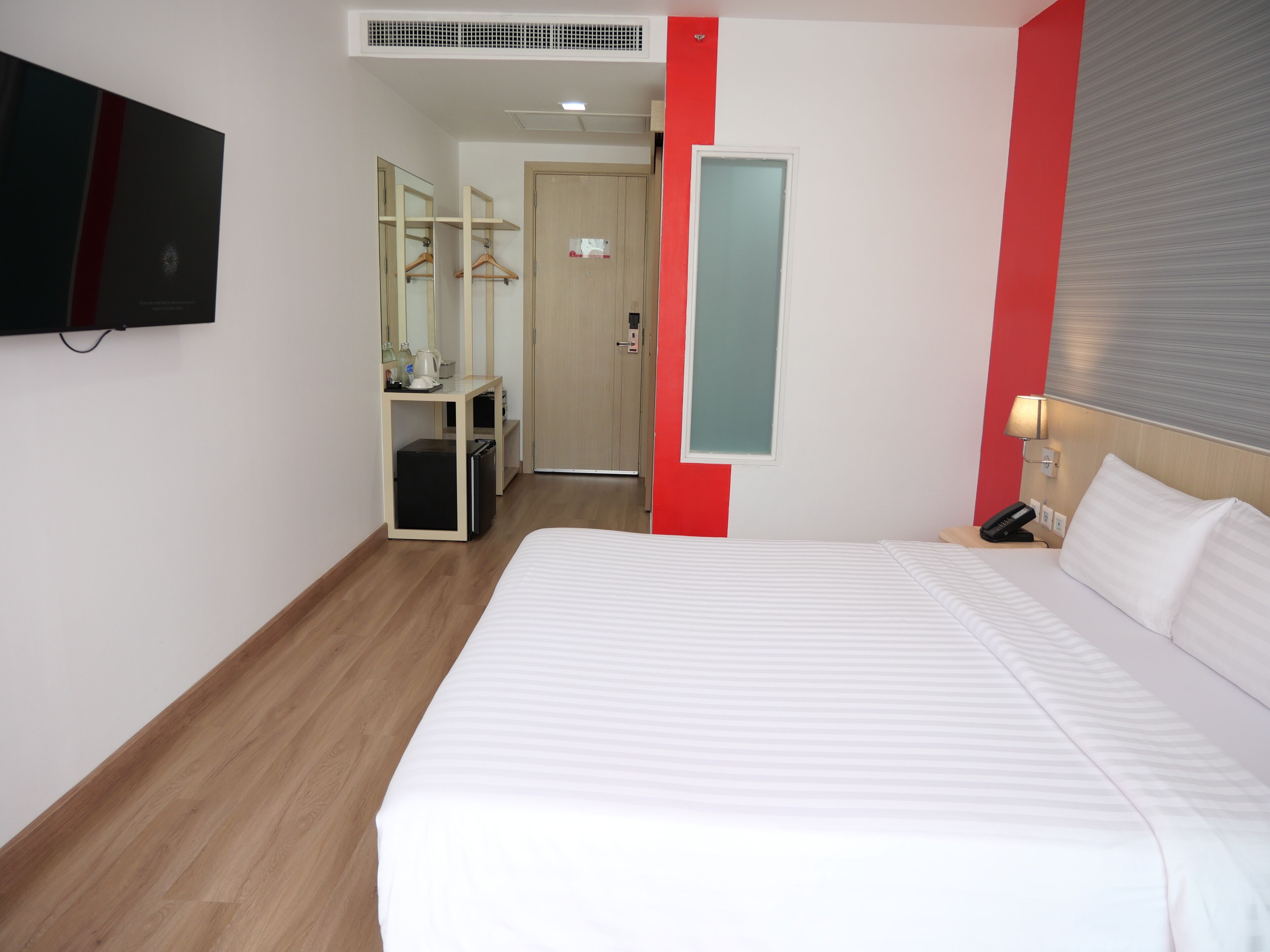 A hotel room with a large bed, a desk with a computer, a mirror, a wardrobe, and a TV mounted on the wall.