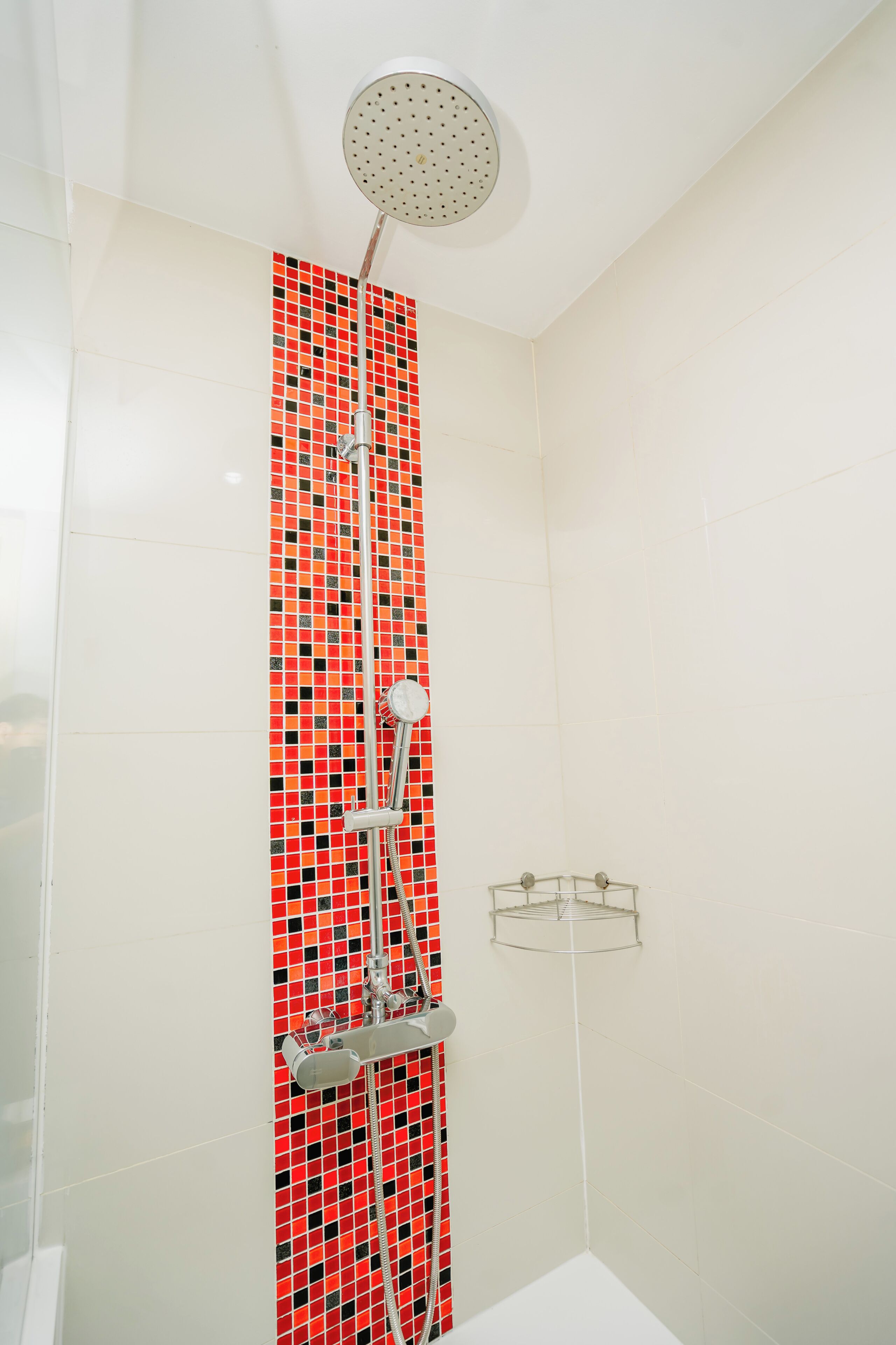 A shower with a red and black mosaic tile wall, a ceiling-mounted showerhead, and a clear glass shelf.