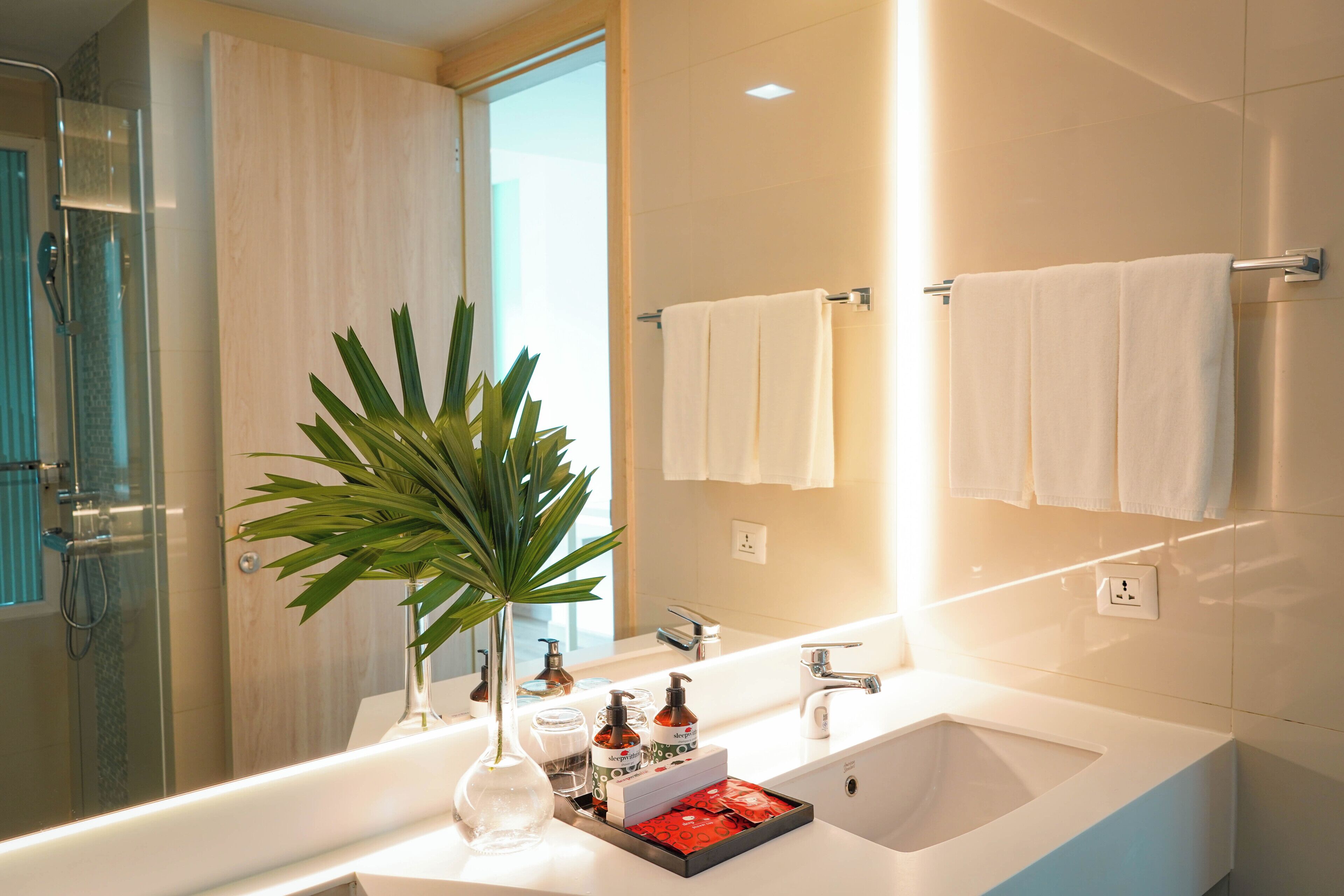 Une salle de bain moderne avec une douche à parois de verre, un grand miroir et un lavabo blanc agrémenté d’articles de toilette.