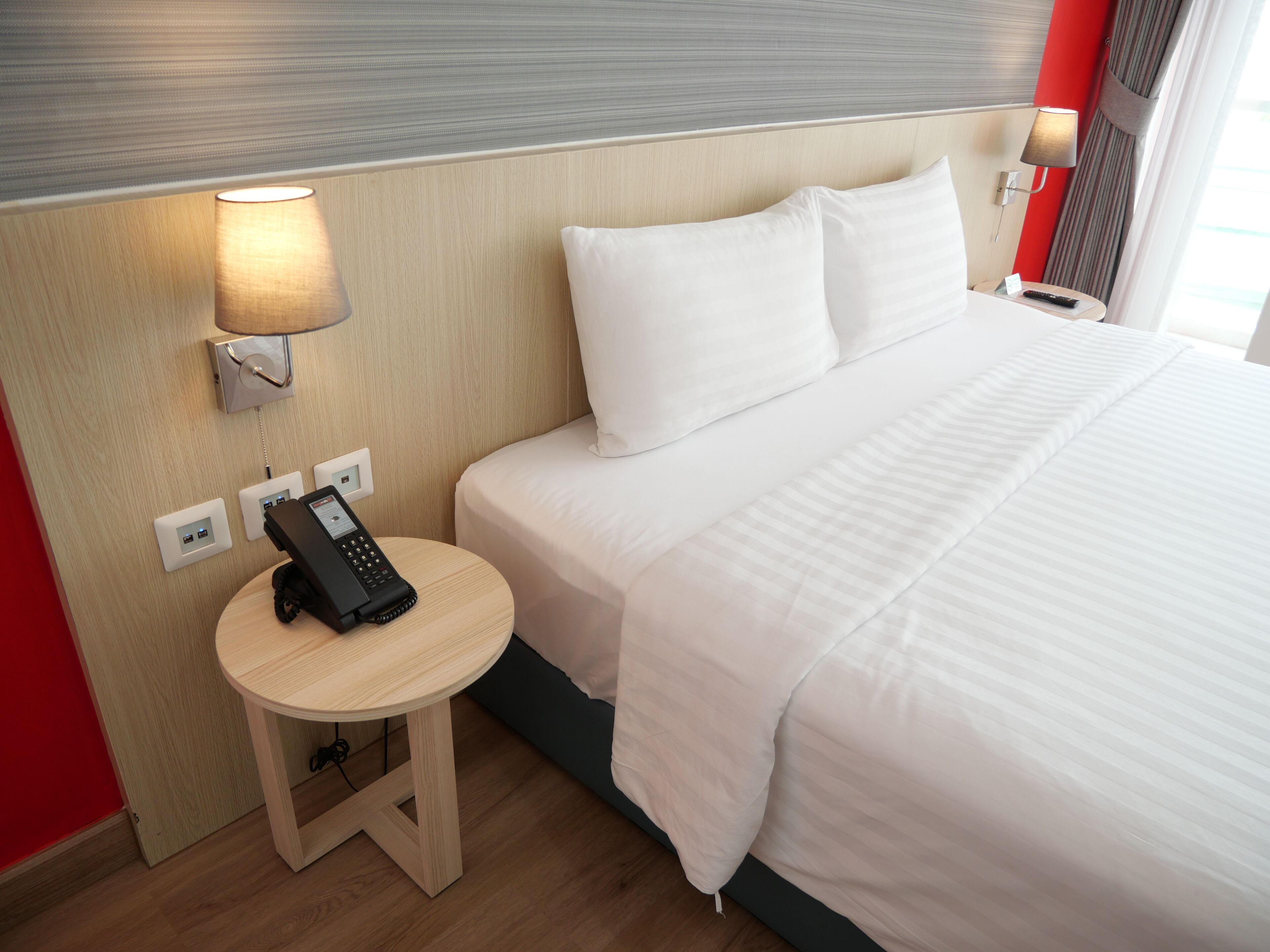 A hotel room with a bed, a bedside table with a phone, a lamp, and a wall-mounted control panel.