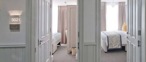Una habitación de hotel con una cama, una mesita de noche, una silla y una pequeña mesa. Se observa una puerta con una placa de número y una lámpara sobre ella.