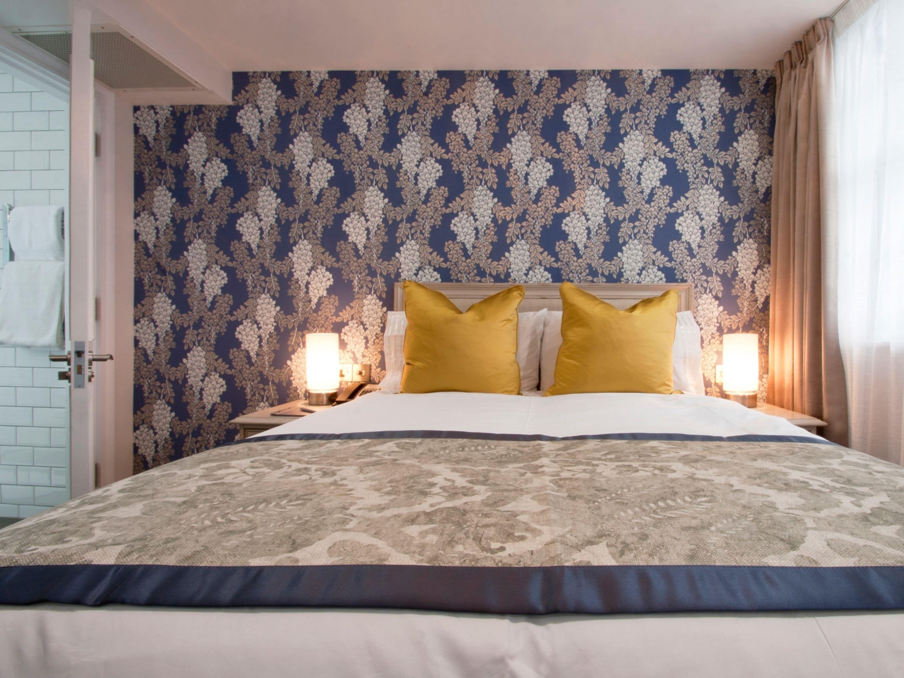 A hotel room with a patterned wallpaper, a bed with a patterned bedspread, two bedside lamps, and a nightstand with a phone and a lamp.