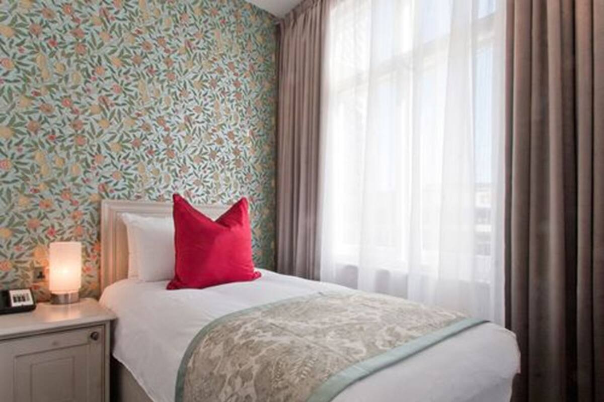 A hotel room with a floral wallpaper, a bed with a red pillow, a nightstand with a lamp, and sheer curtains.
