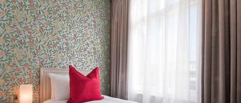 A hotel room with a floral wallpaper, a bed with a red pillow, a nightstand with a lamp, and sheer curtains.