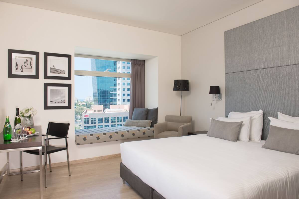 Une chambre d’hôtel moderne dotée d’un grand lit, d’un coin salon avec vue, d’un bureau avec une chaise et de tableaux encadrés au mur.