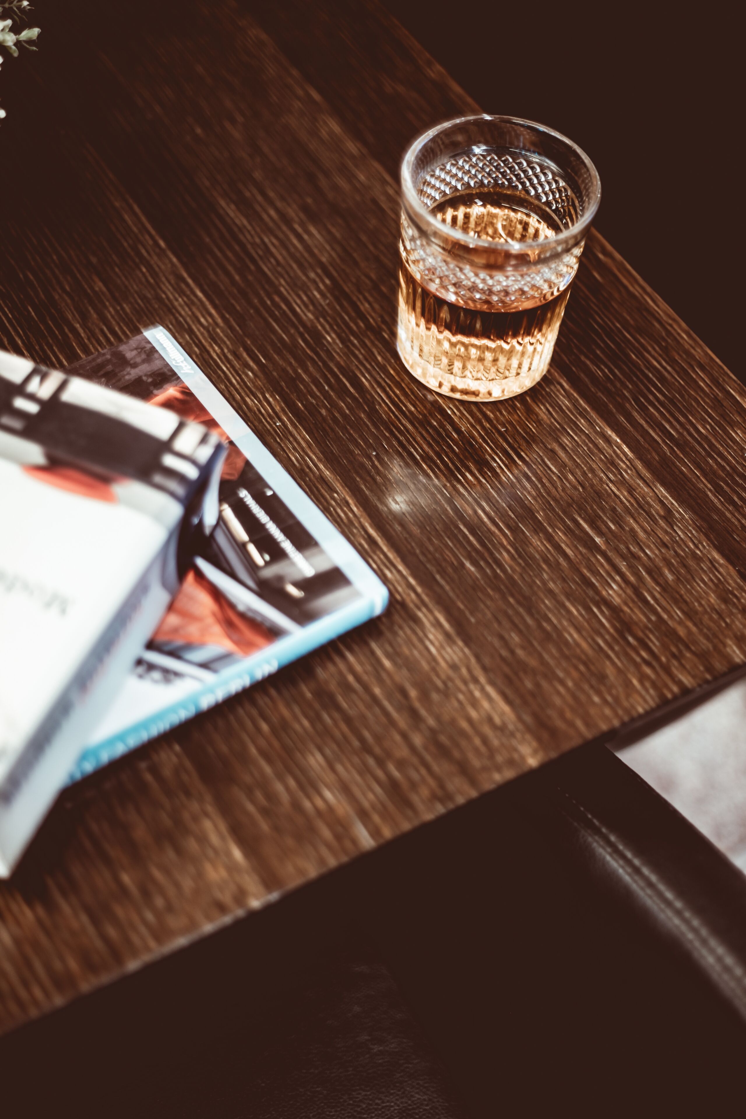 Una mesa de madera con un vaso de líquido ámbar, una baraja de cartas y un libro.