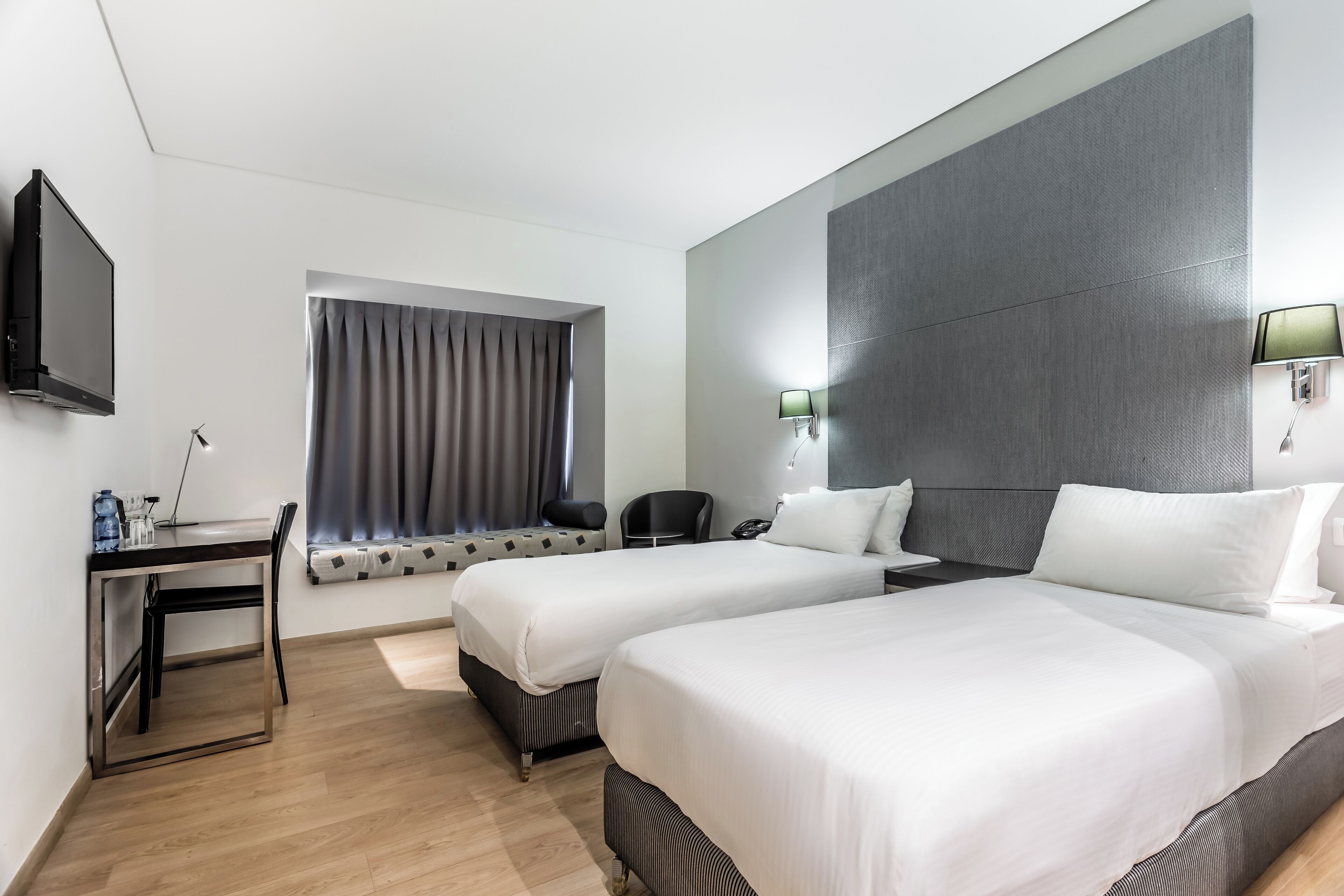 Quarto de hotel com duas camas, uma escrivaninha, uma cadeira, uma televisão e uma janela com cortinas.