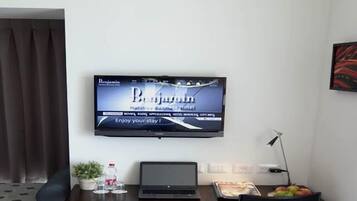 Une chambre d’hôtel moderne avec un téléviseur à écran plat fixé au mur, sur lequel est affiché le nom de l’hôtel. On y trouve un bureau avec un ordinateur portable, une chaise et un plateau de nourriture. Une petite plante en pot et une bouteille d’eau sont posées sur le bureau.