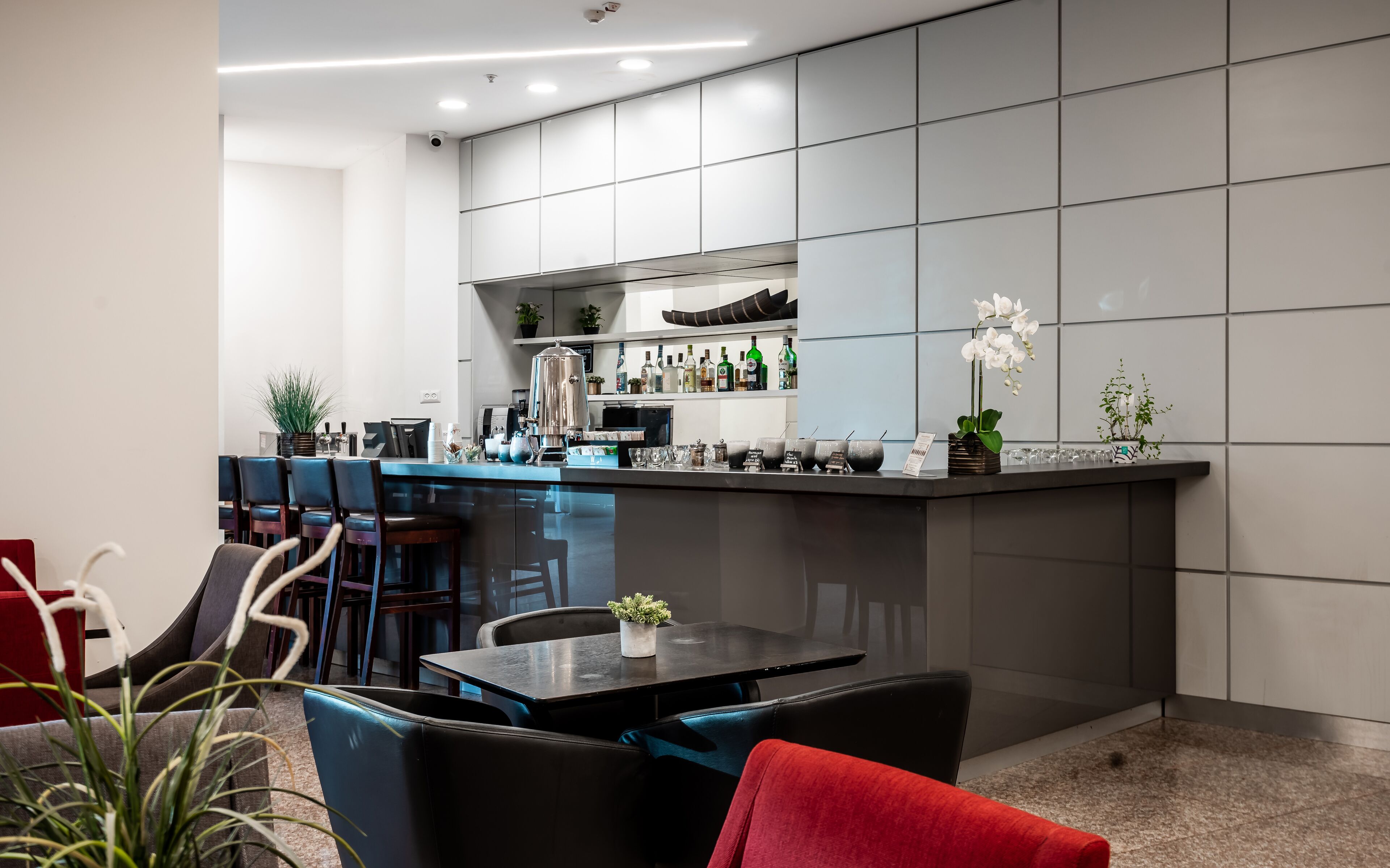 A modern kitchen with a bar area, featuring a countertop with various bottles and a coffee machine. There are chairs and a table in the dining area.