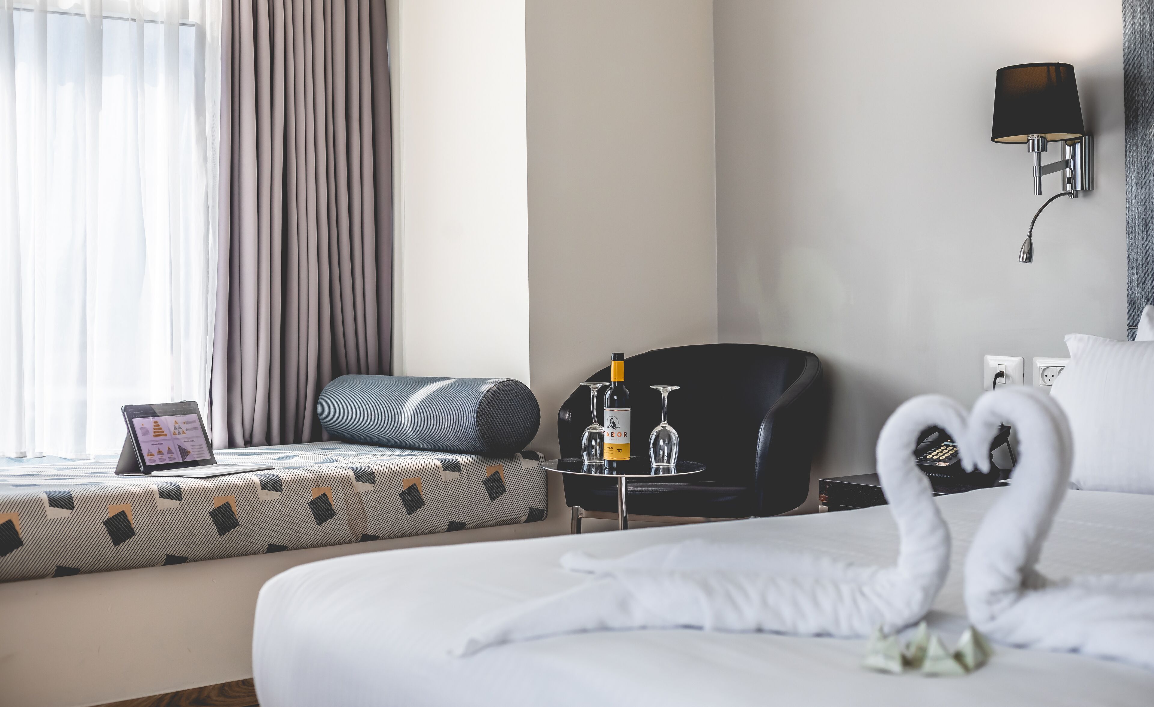 Une chambre d’hôtel avec un lit soigneusement fait, un fauteuil noir, une petite table avec une bouteille et des verres, et une tablette posée sur le lit.