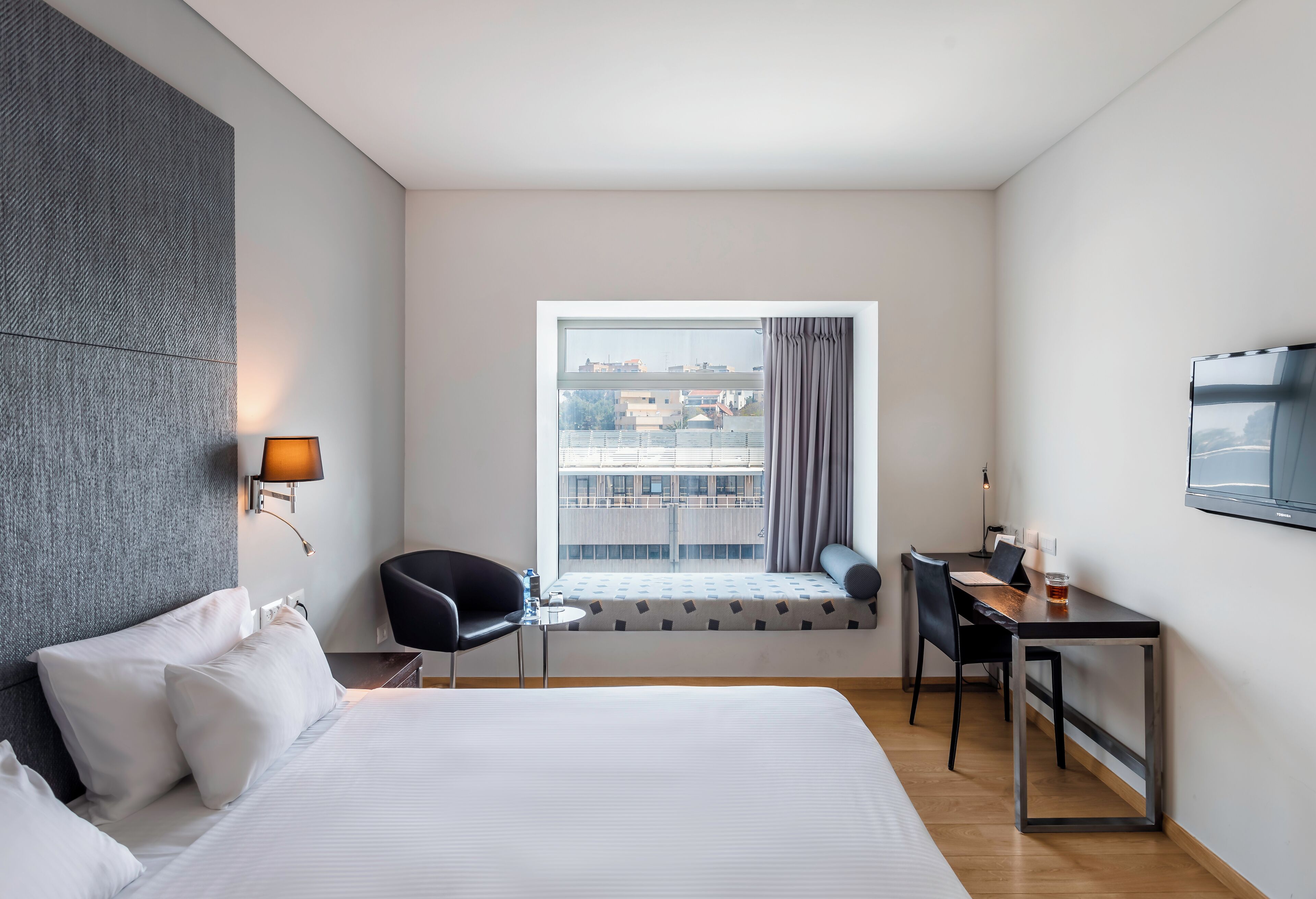 A modern hotel room with a large bed, a desk, a chair, a window with a view of buildings, and a mounted TV.