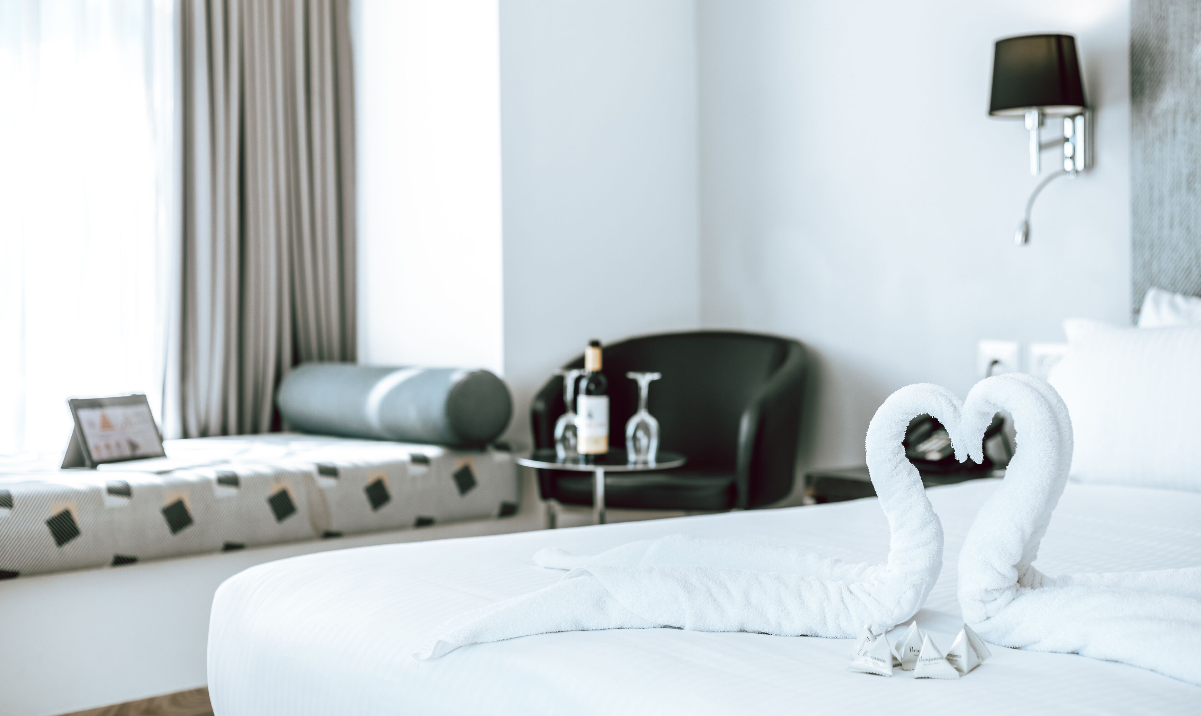 A hotel room with a neatly made bed, a black armchair, a small table with a bottle and glasses, and a wall-mounted lamp.