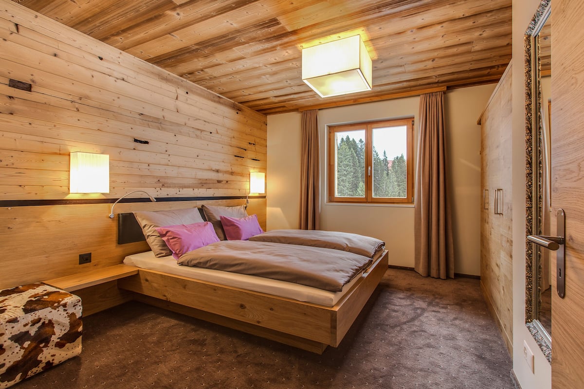 A wooden cabin bedroom with a bed, bedside lamps, a window with curtains, and a door.