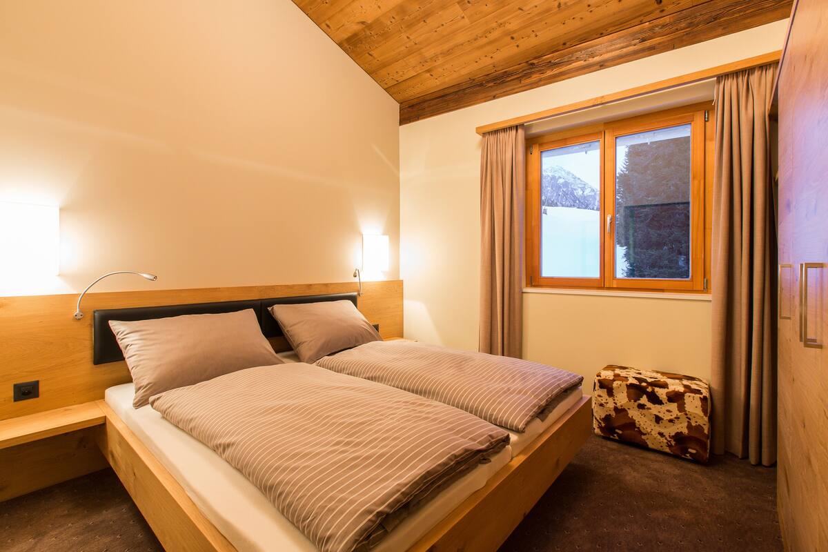 A bedroom with a wooden headboard, a bed with striped bedding, a bedside table, a suitcase, and a window with curtains.