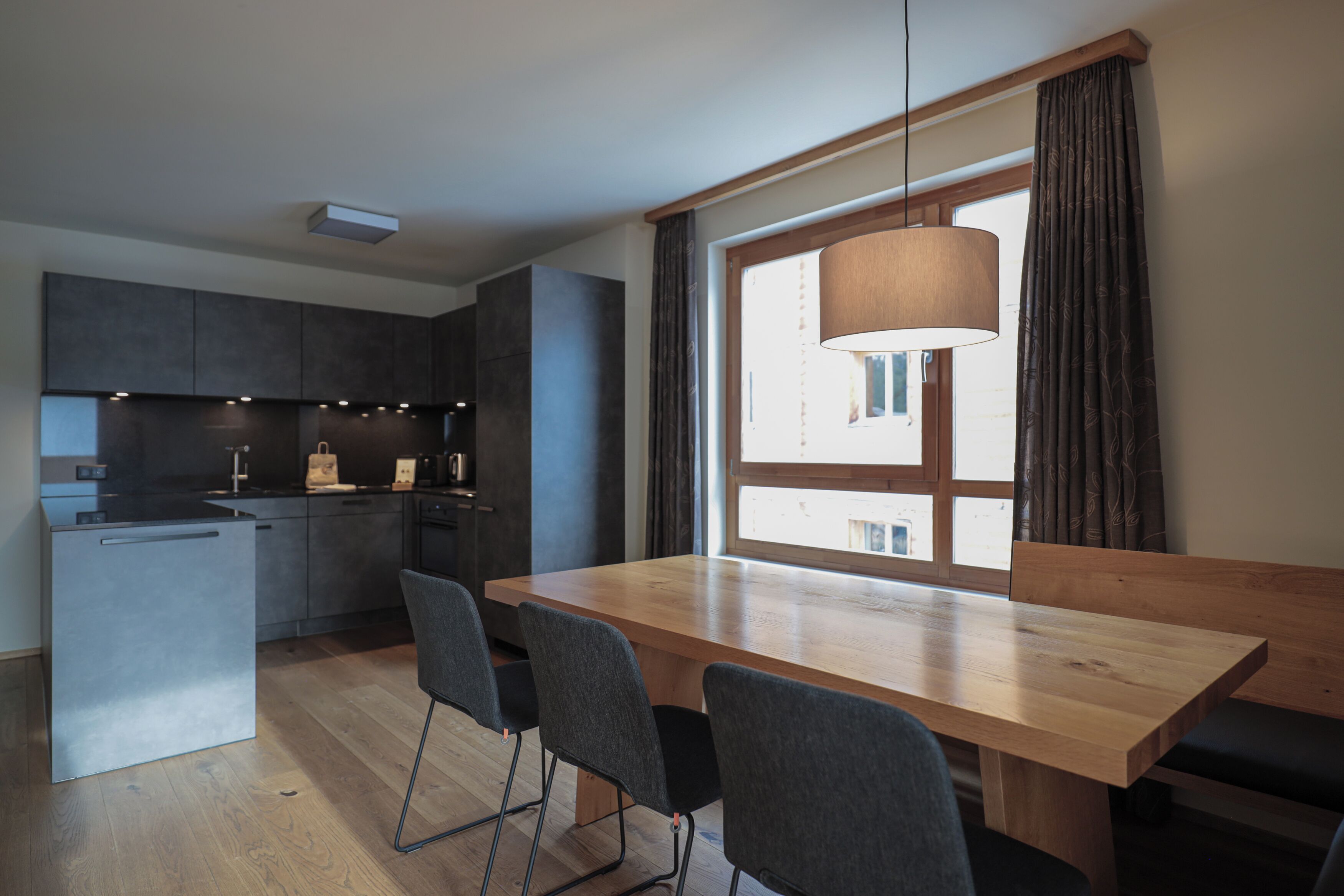 Uma cozinha moderna com armários escuros, uma mesa de jantar de madeira e uma janela grande com cortinas.