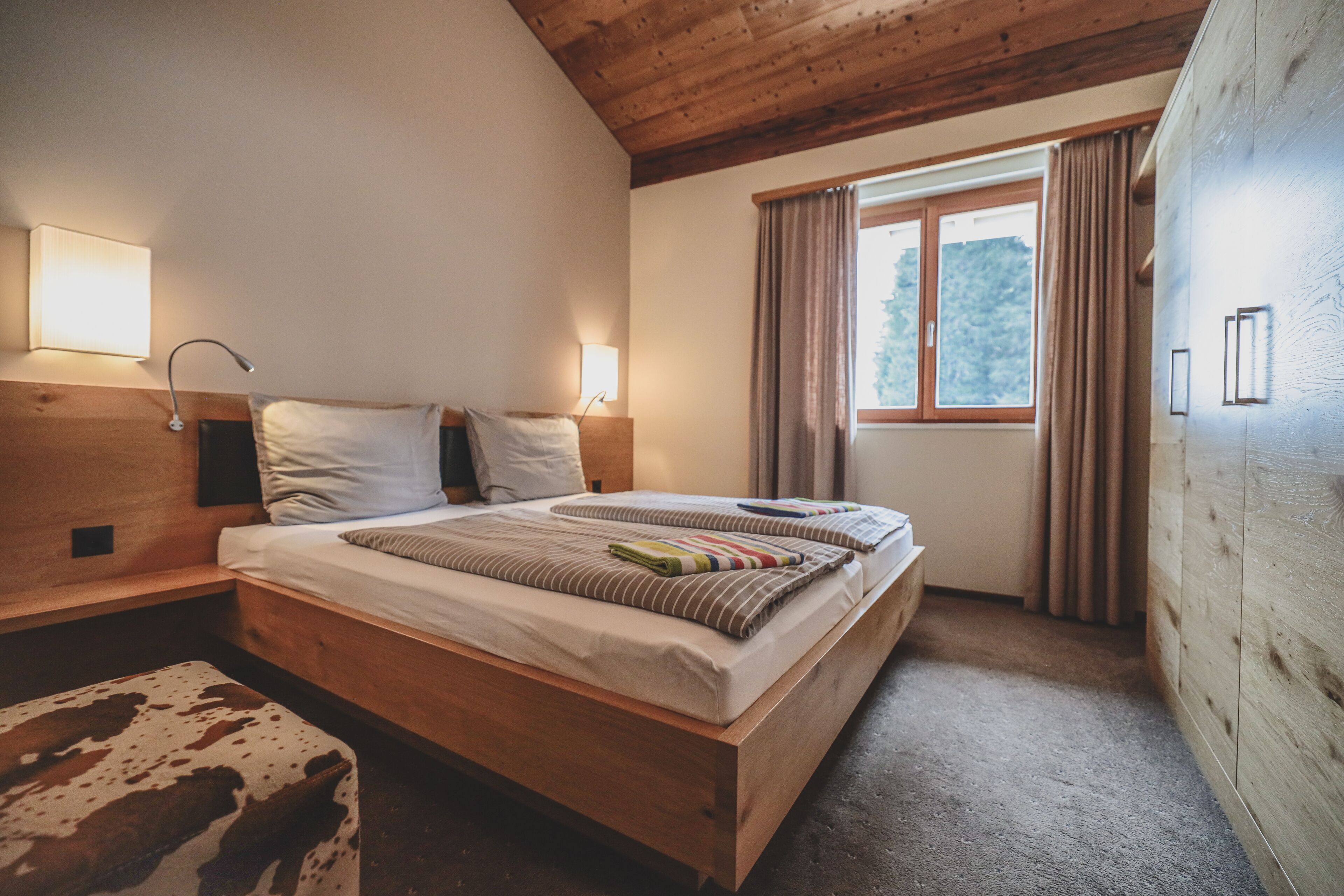 Un dormitorio acogedor con una cama de madera, dos mesitas de noche, una mesita auxiliar, una ventana con cortinas y un cabecero de madera.