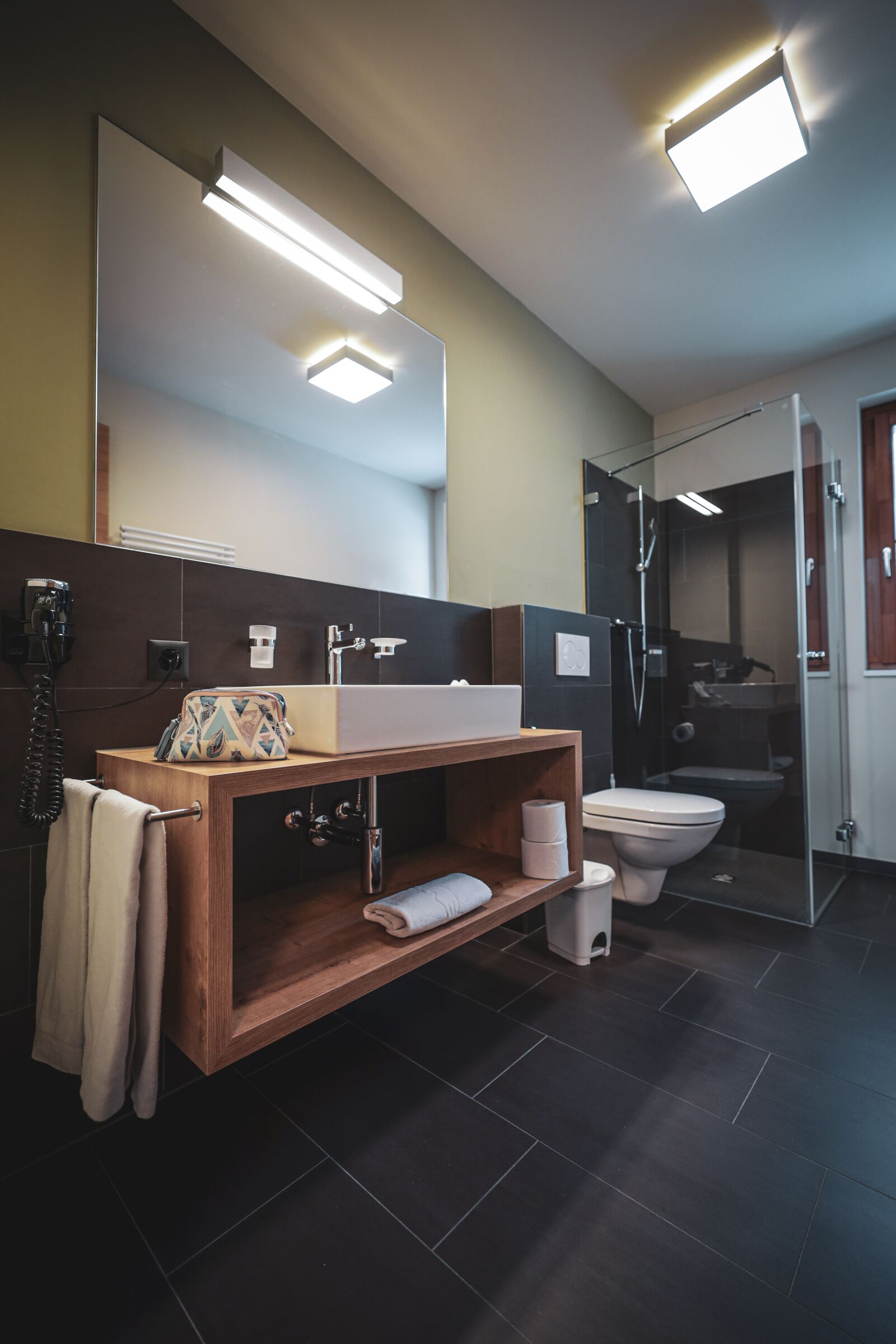 A modern bathroom with a wooden vanity, a large mirror, a glass shower enclosure, and a toilet.