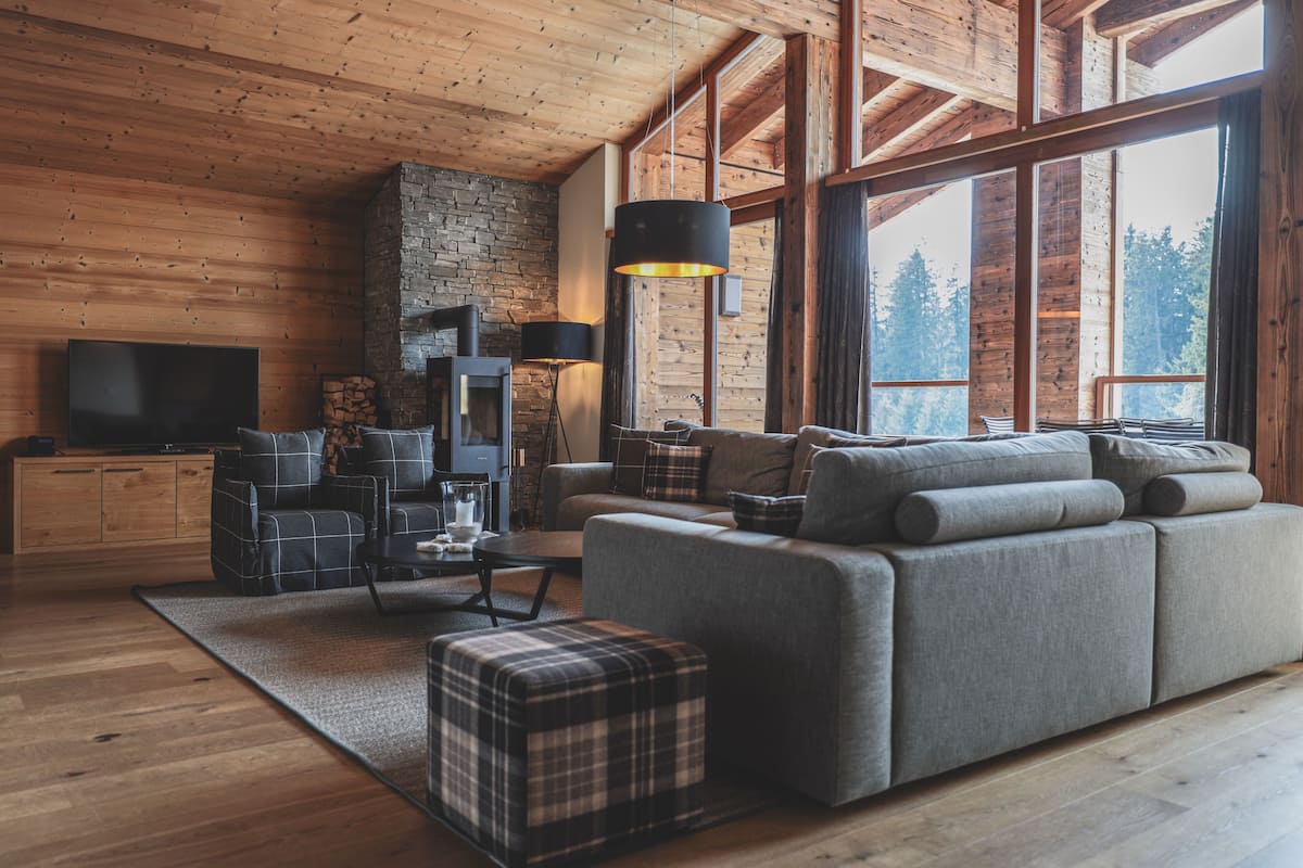 A cozy living room with a large gray sofa, a plaid ottoman, and a flat-screen TV against a wooden wall.