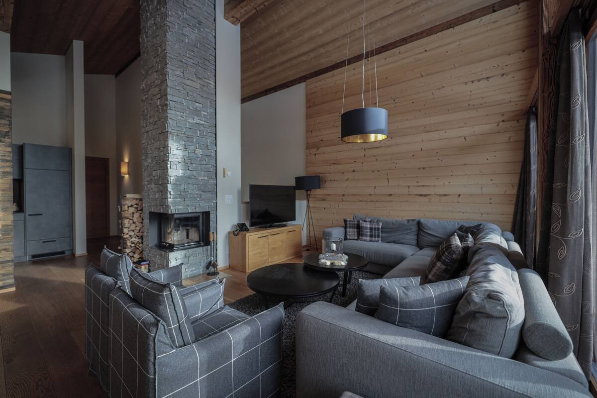 A modern living room with a stone fireplace, a sofa set, a television, and a wooden ceiling.