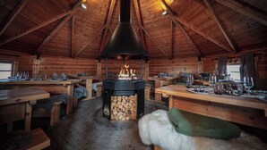 Interior de una cabaña de madera con chimenea central, mesas de madera, bancos y un banco cubierto de pieles.