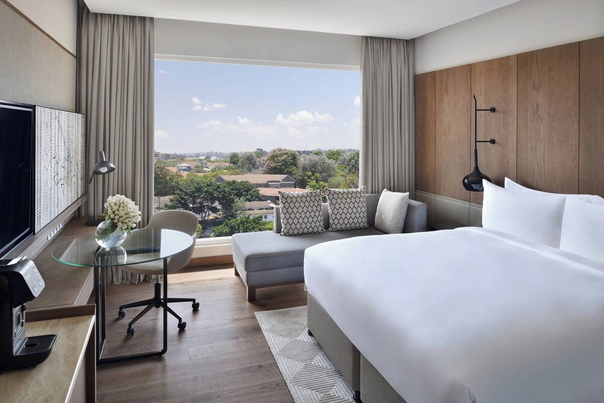 A modern hotel room with a large bed, a desk with a glass top, a chair, and a view of the cityscape.