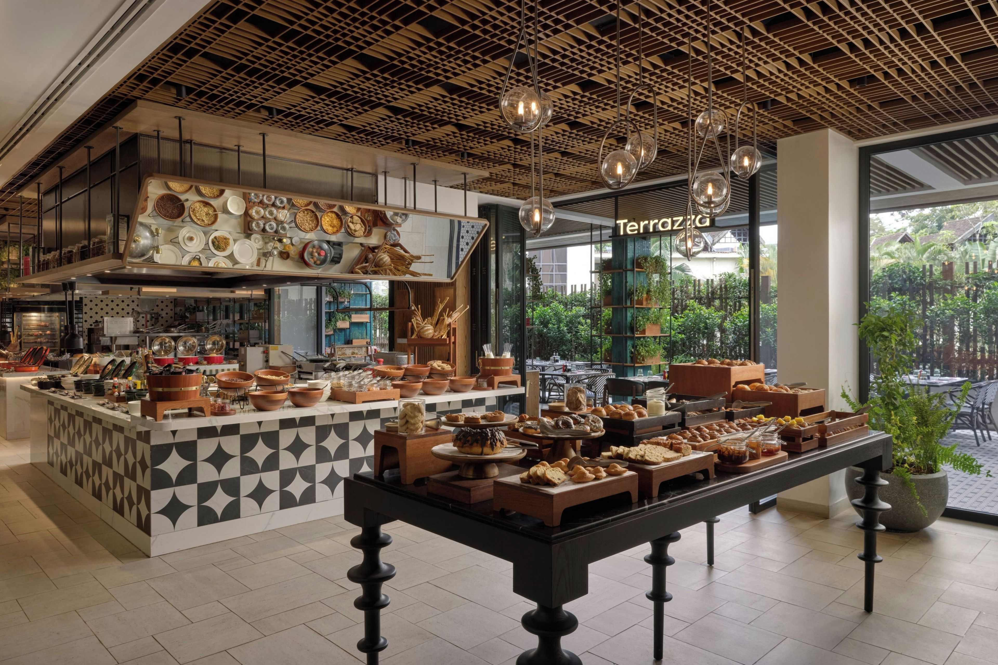 A modern cafe interior with a central buffet table, a kitchen area, and a view of the outdoors.