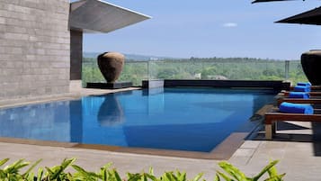 A modern outdoor swimming pool with clear blue water, surrounded by lounge chairs and umbrellas, and a view of a forested landscape in the background.