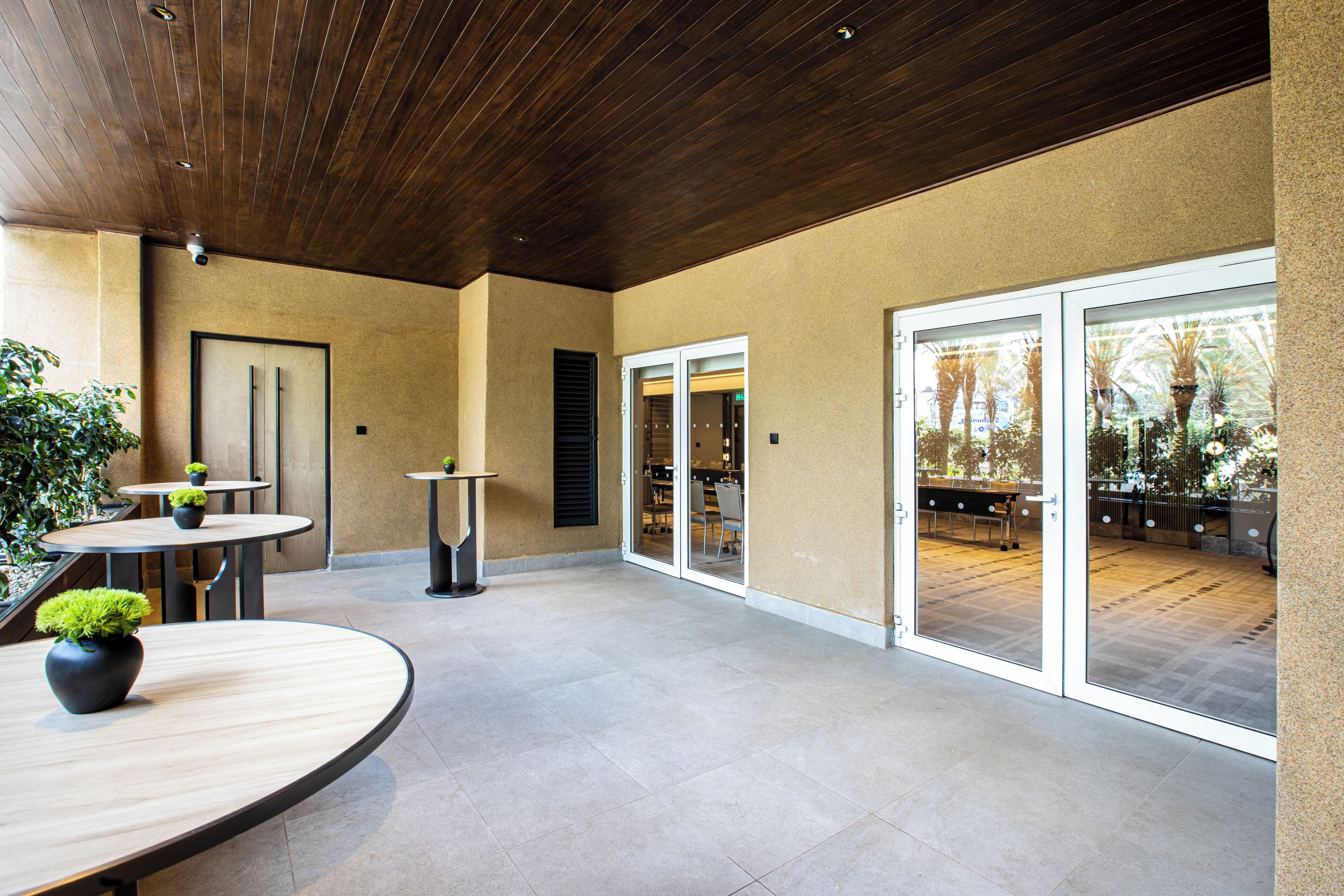 Ein überdachter Patio-Bereich mit mehreren Tischen und Stühlen, einer Holzbalkendecke und einer Glastür, die auf eine Terrassenfläche führt.