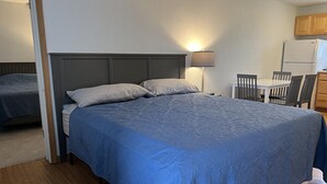 Ein Schlafzimmer mit Bett, Nachttisch, Lampe und einer Küchenzeile inklusive Kühlschrank und Stühlen.