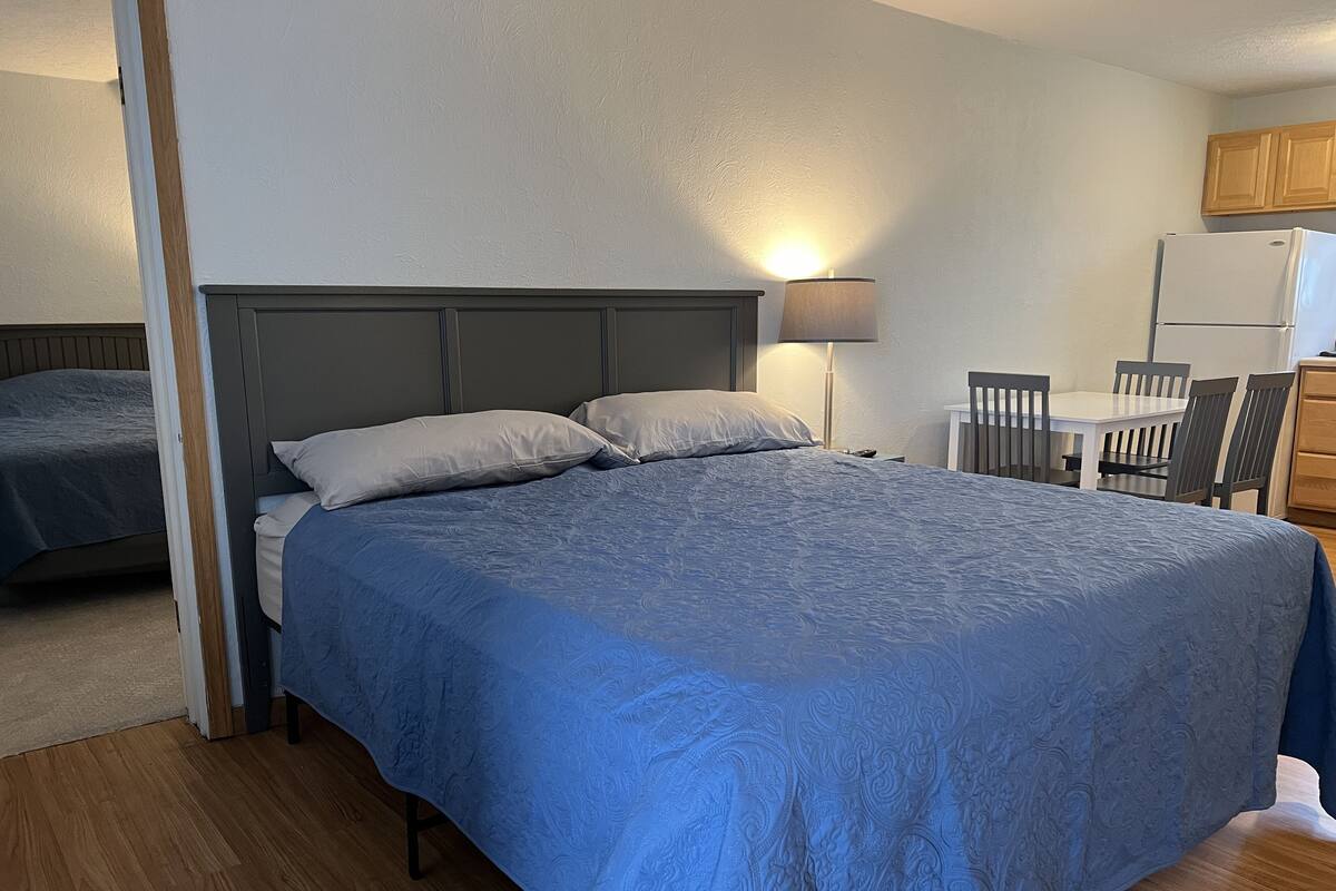 Habitació amb llit, taula de nit, làmpada i una zona de cuina compacta amb nevera i cadires.