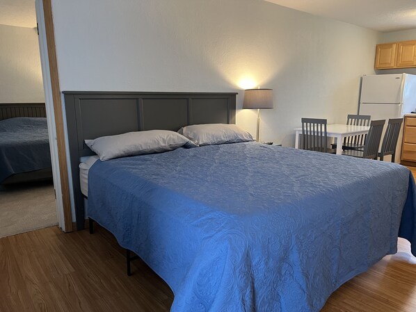 A bedroom with a bed, bedside table, lamp, and a kitchenette area with a refrigerator and chairs.