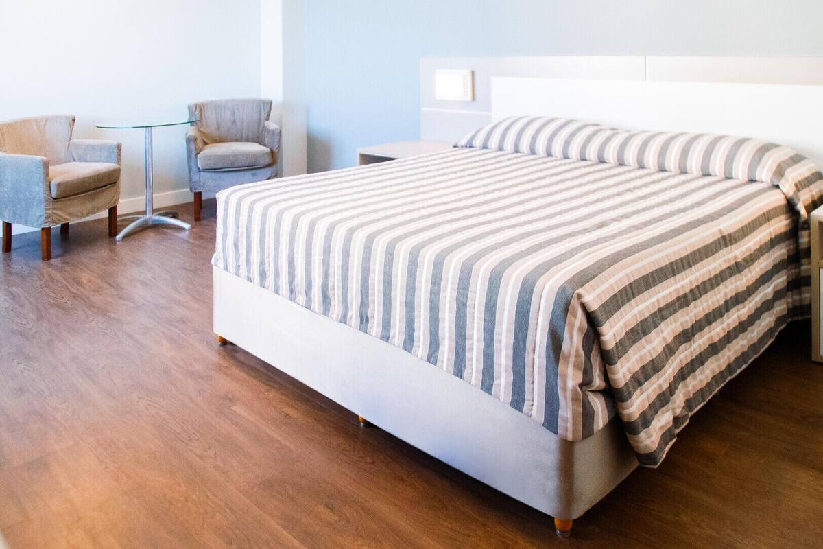 A hotel room with a bed, two chairs, a nightstand, and a wooden floor.