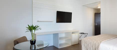 A modern hotel room with a bed, a desk with a flat-screen TV, a glass table, two chairs, and a potted plant.