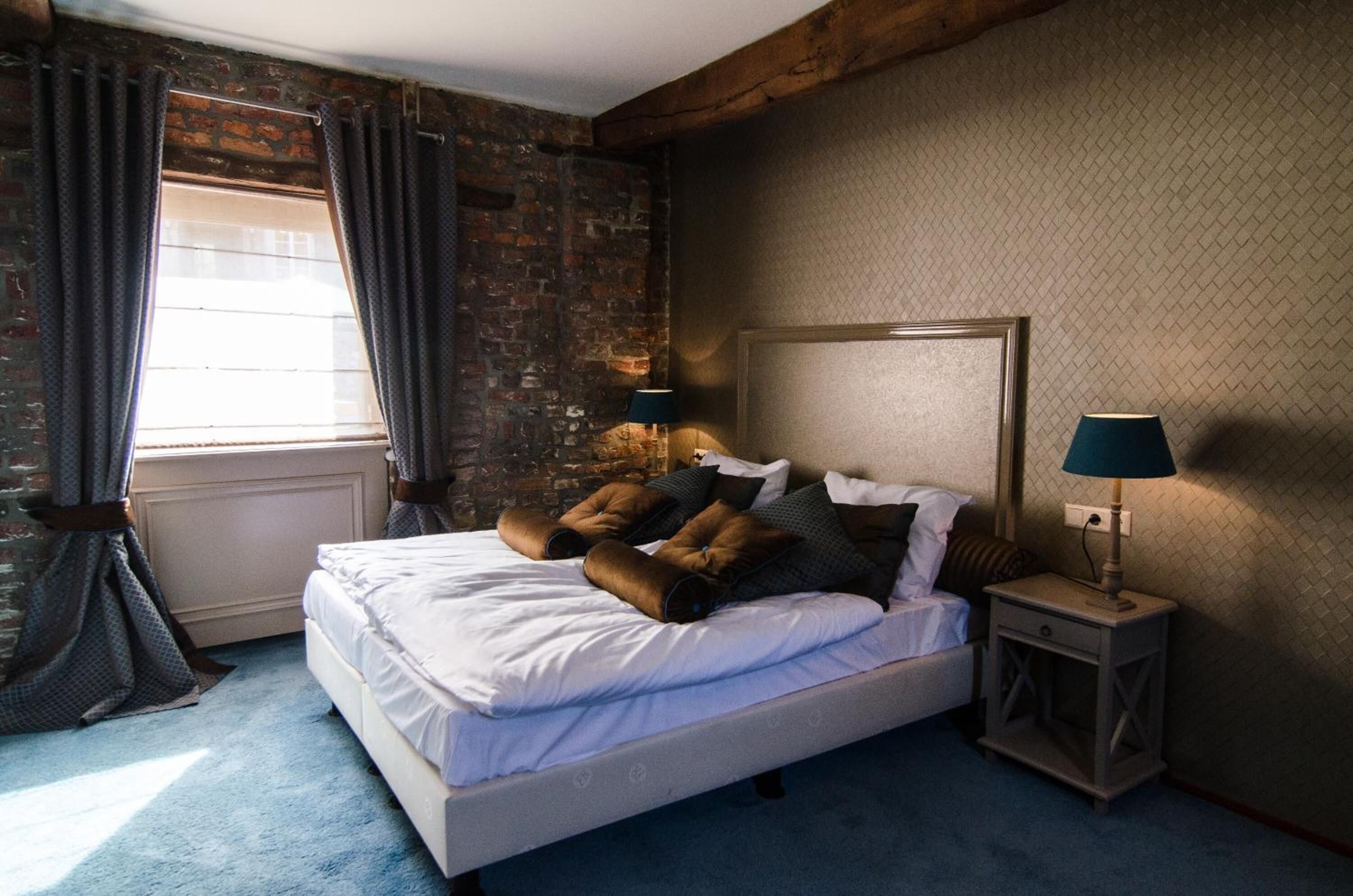 A bedroom with a brick wall, a bed with white linens, two bedside lamps, and a window with curtains.