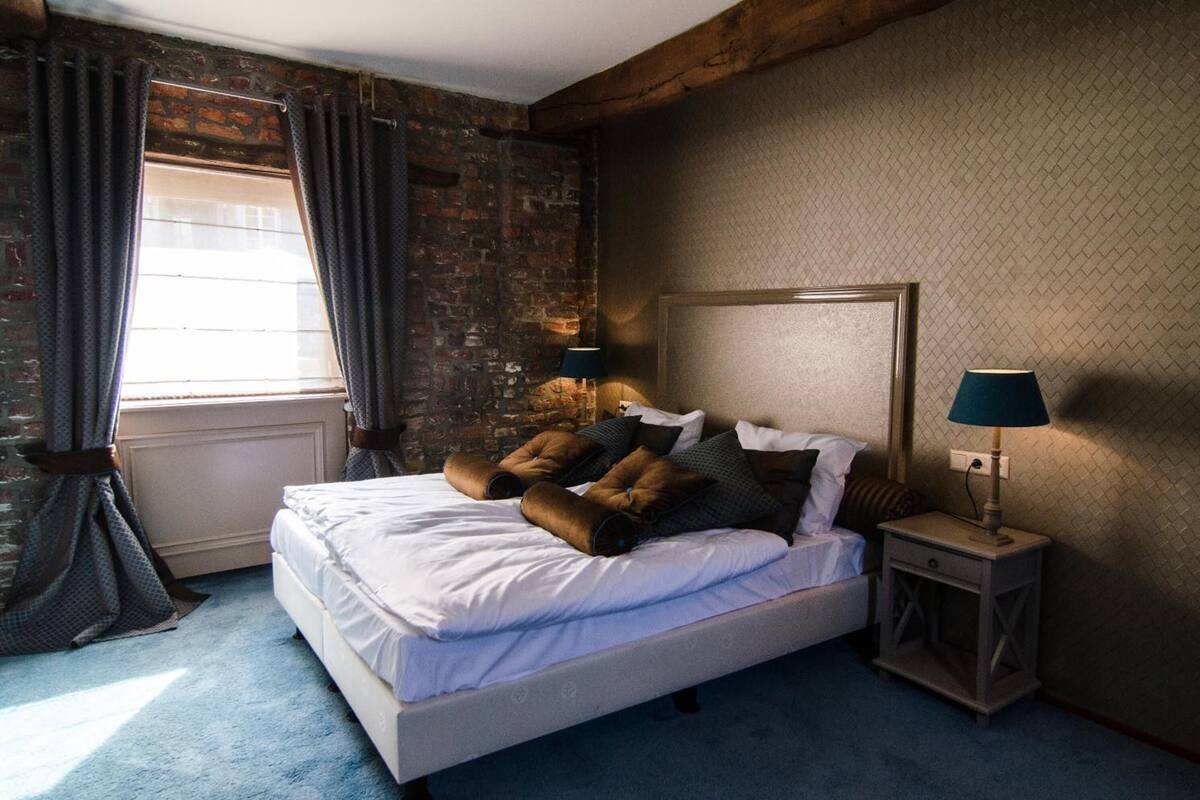A bedroom with a brick wall, a bed with white linens, two bedside lamps, and a window with curtains.