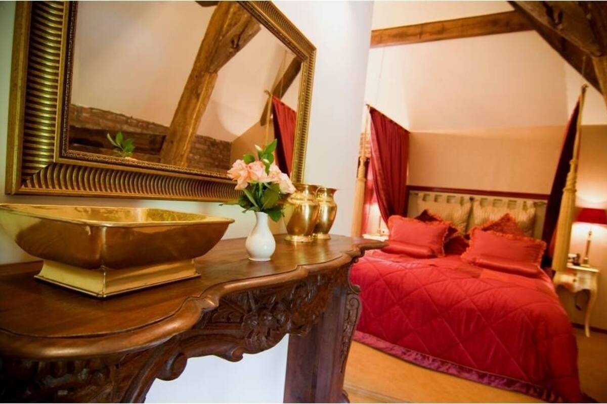 A bedroom with a wooden dresser, a large mirror, a red bedspread, and a vase with flowers.