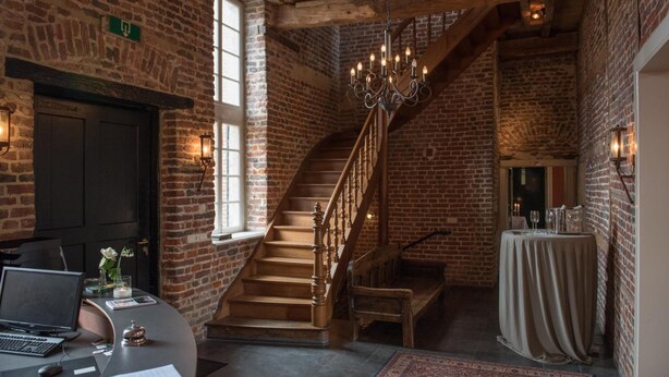 A rustic interior with exposed brick walls, a wooden staircase, a chandelier, and a round table with a tablecloth.