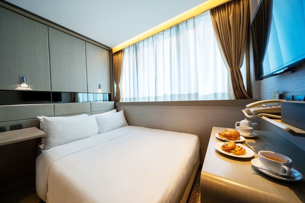 A hotel room with a large bed, a bedside table with a cup of coffee and pastries, a telephone, and a television on the wall.