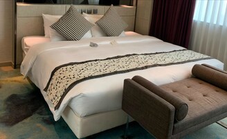 Una cama cuidadosamente tendida, con un plaid estampado, dos almohadas blanco y negro, y un sillón marrón en una habitación de hotel moderna.