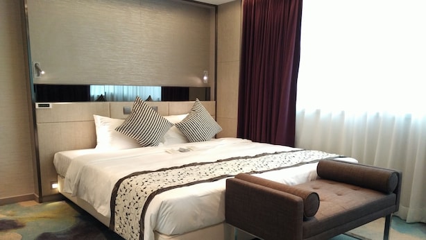 Ein Hotelzimmer mit einem großen Bett, einem braunen Sessel, einem Kopfteil mit einer hellen Platte und einem Fenster mit dunklen Vorhängen.