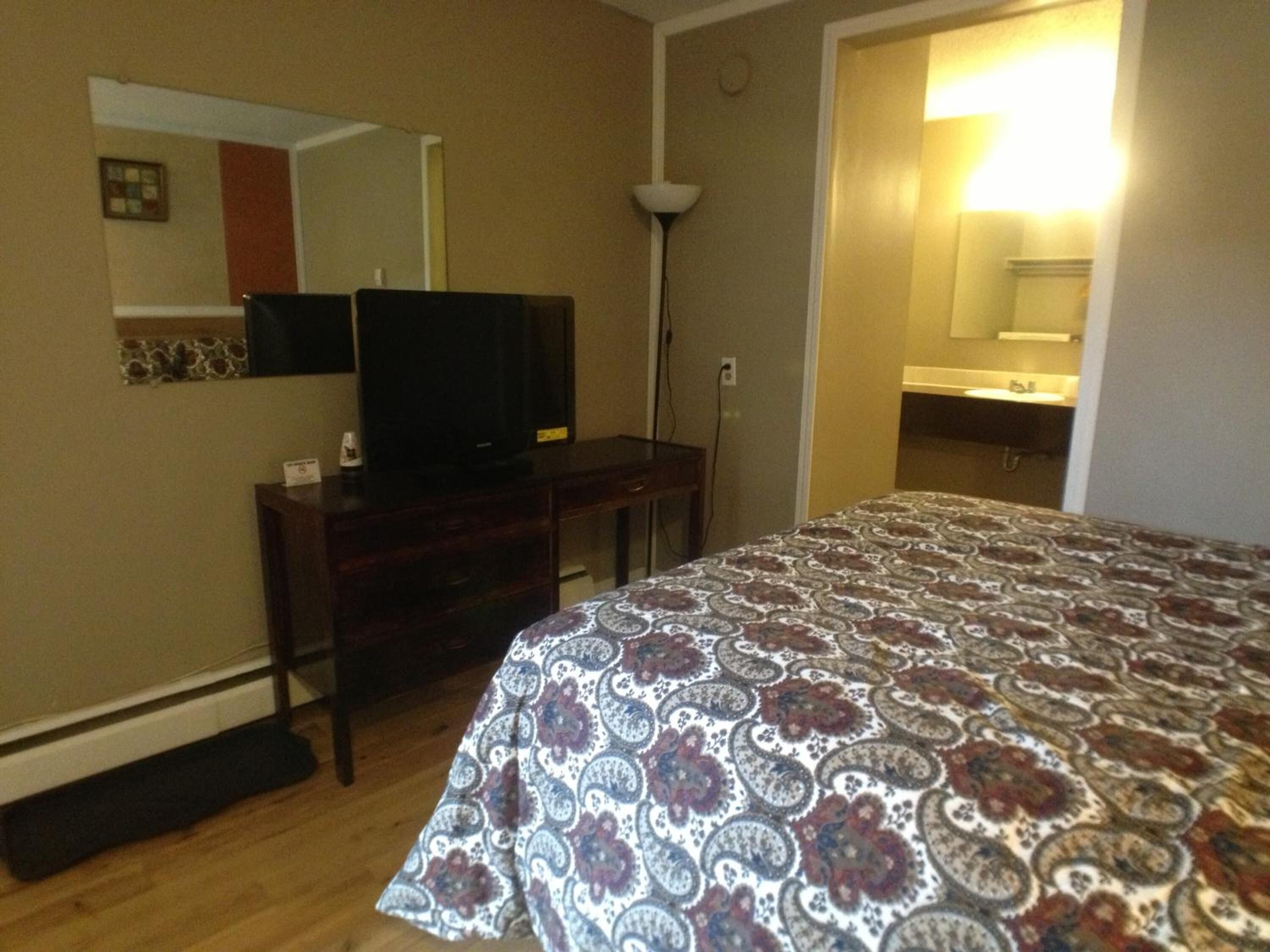 Una habitación de hotel con una cama, un televisor sobre un aparador de madera, un espejo y un baño a la vista a través de una puerta abierta.