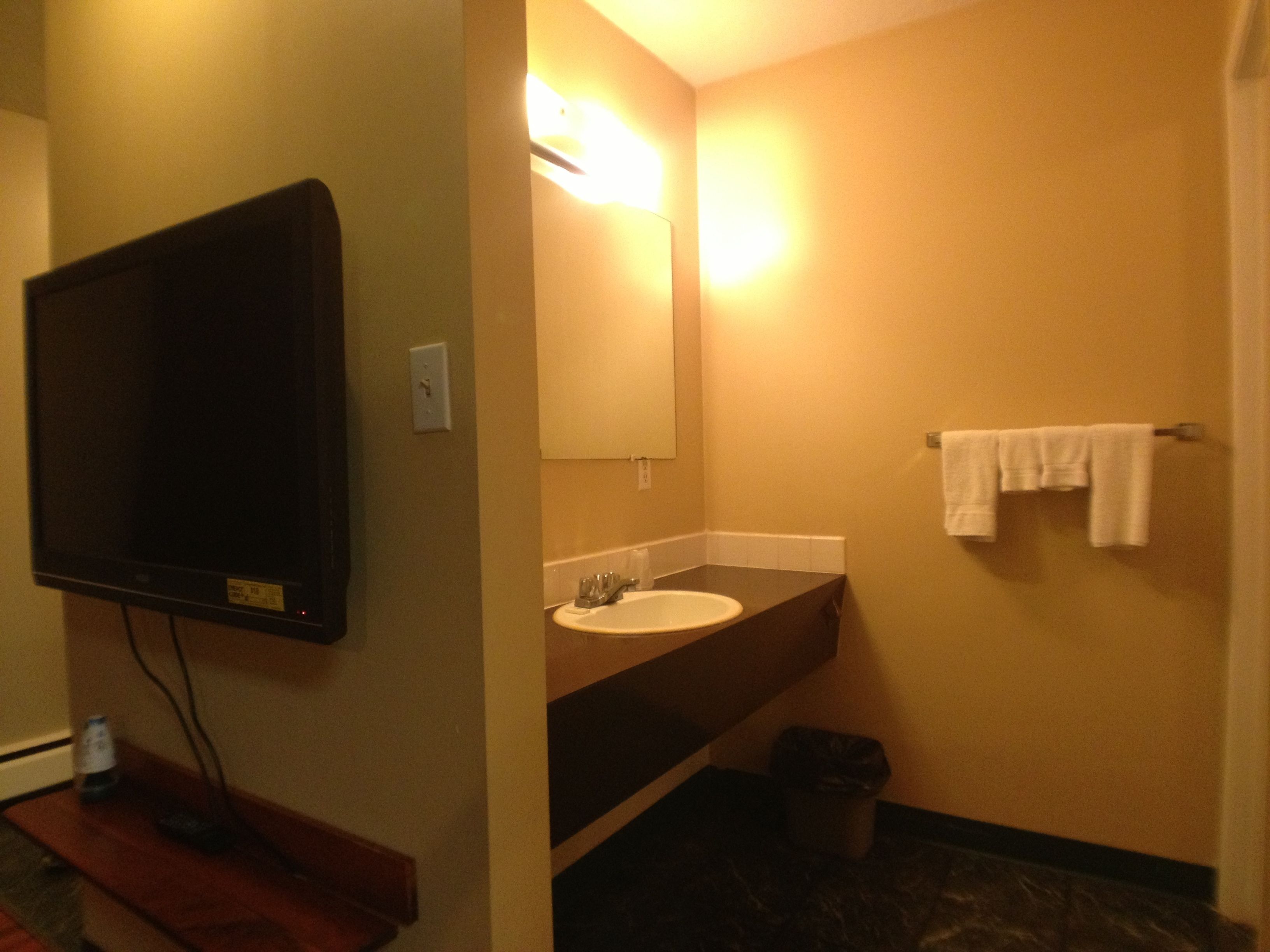 A bathroom with a flat-screen TV mounted on the wall, a sink with a mirror, and a towel rack with towels.