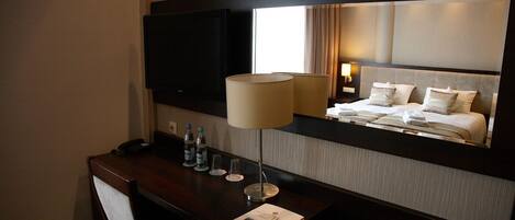 A hotel room with a large bed, a desk with a lamp, a TV, and a mirror reflecting the bed.