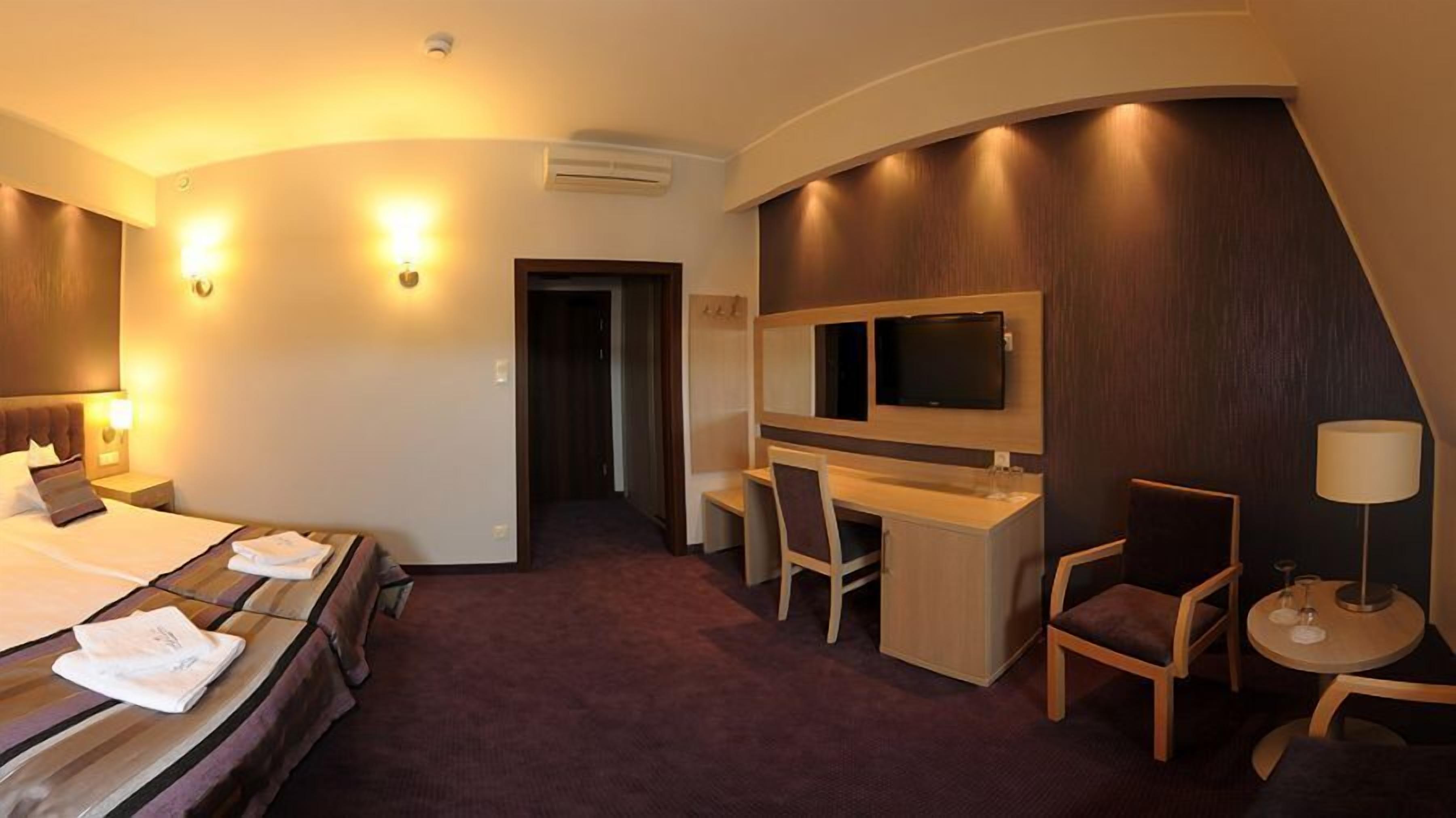 A hotel room with a bed, desk, TV, and seating area.